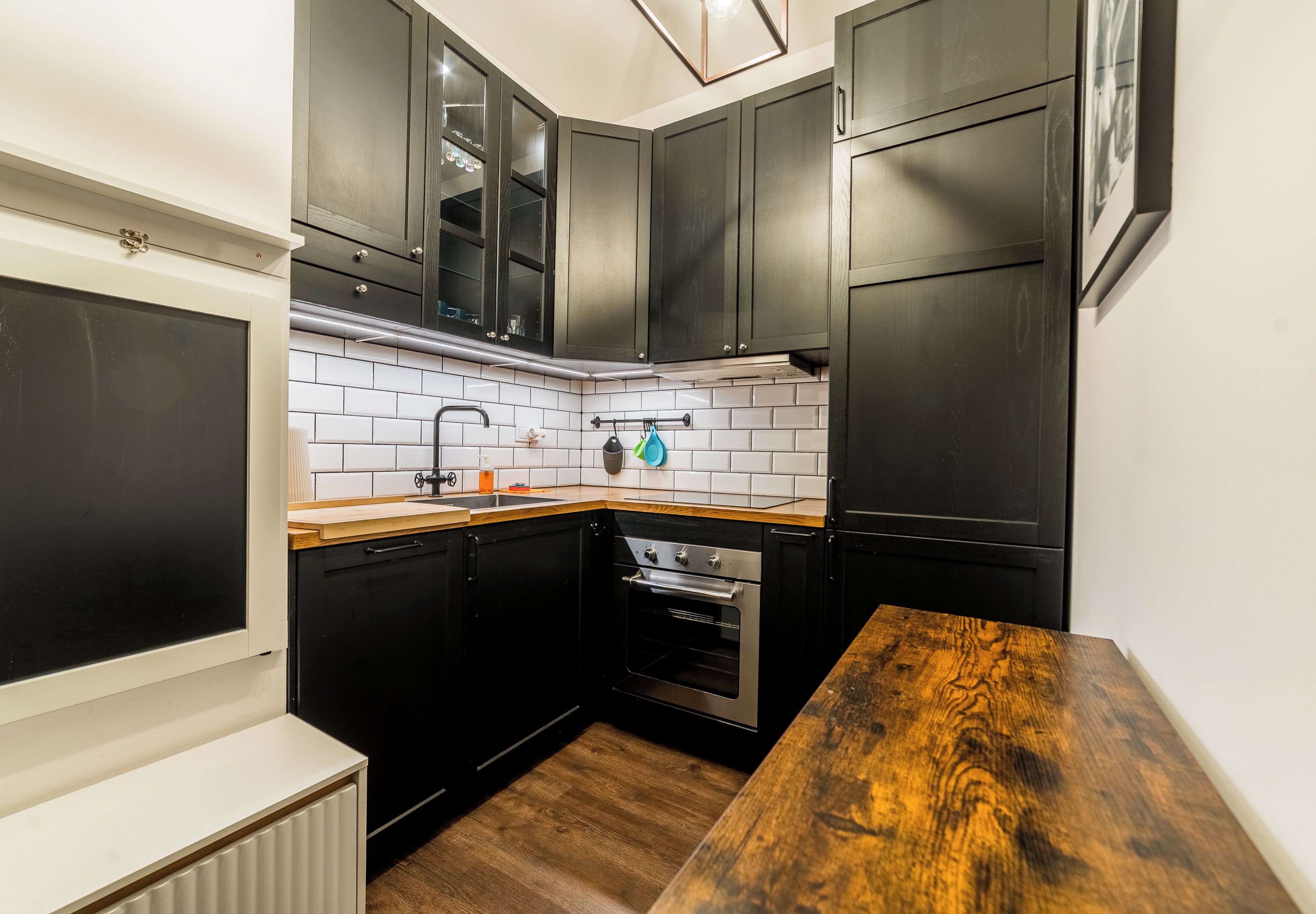 Une cuisine de taille réduite avec des meubles de couleur sombre, un crédence en carreaux blancs et un plan de travail en bois.