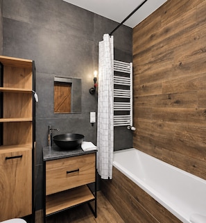 Un baño moderno con tocador de madera, lavabo negro, bañera blanca y cortina de ducha.