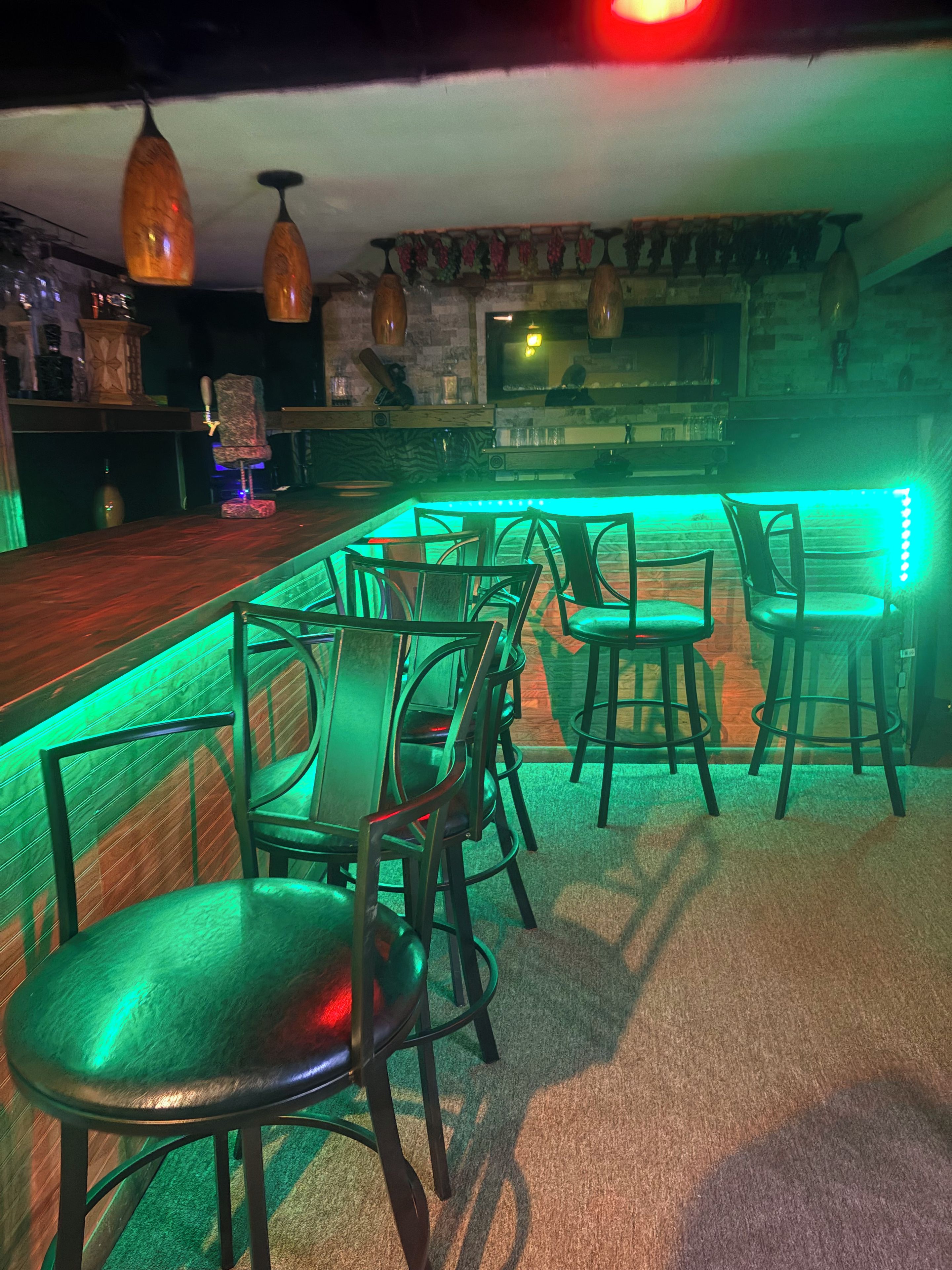 Zona de bar com balcão de madeira, iluminação ambiente verde e vários banquinhos.
