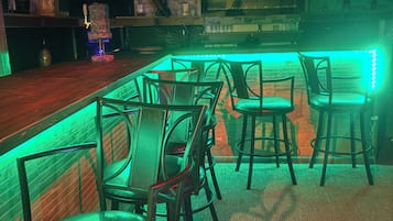 Zona de bar com balcão de madeira, iluminação ambiente verde e vários banquinhos.