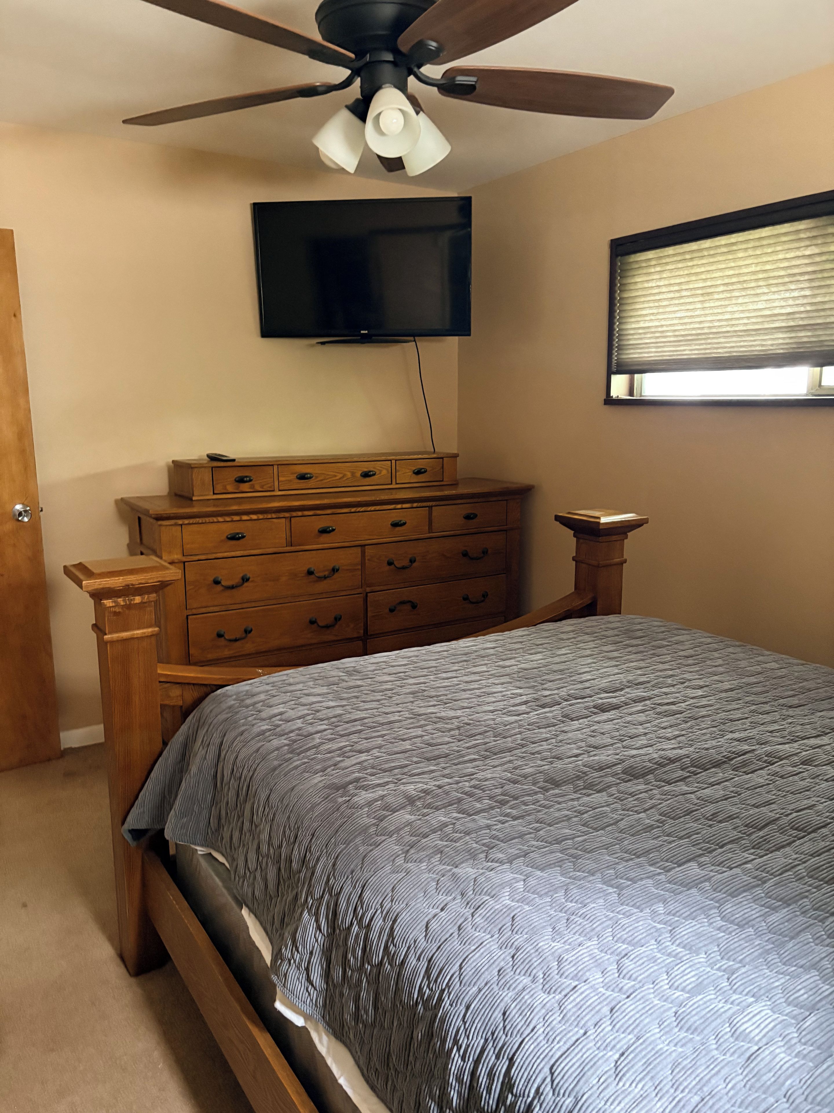 Une chambre à coucher comprenant un lit, une commode en bois, un ventilateur de plafond et un téléviseur fixé au mur.