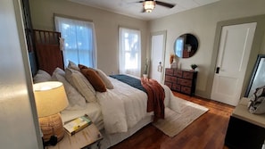 Quarto com cama, mesa de cabeceira, luminária, cômoda, espelho e porta.