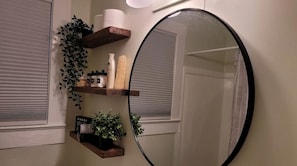 Casa de banho com um espelho grande, prateleiras de madeira com produtos de higiene pessoal e uma planta em vaso.