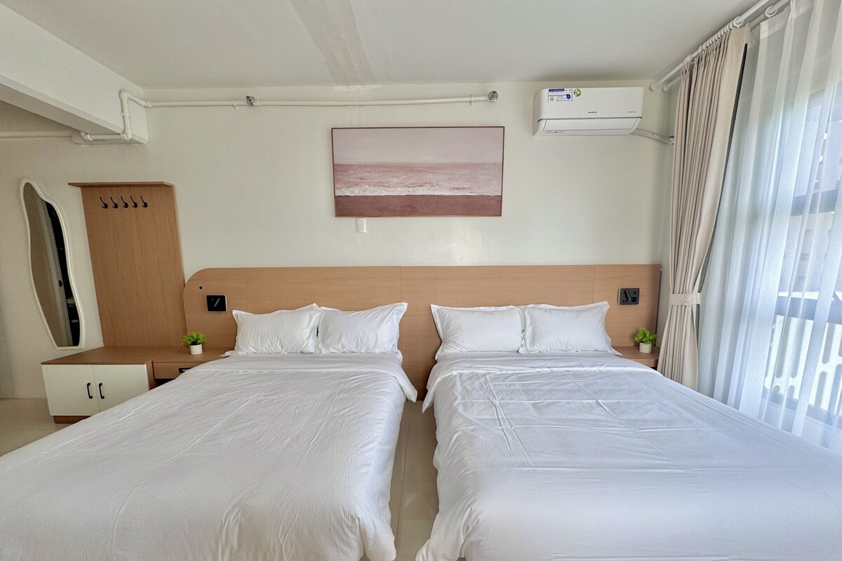 A hotel room with two beds, a wooden headboard, a small bedside table with a plant, a wall-mounted air conditioner, and a window with curtains.