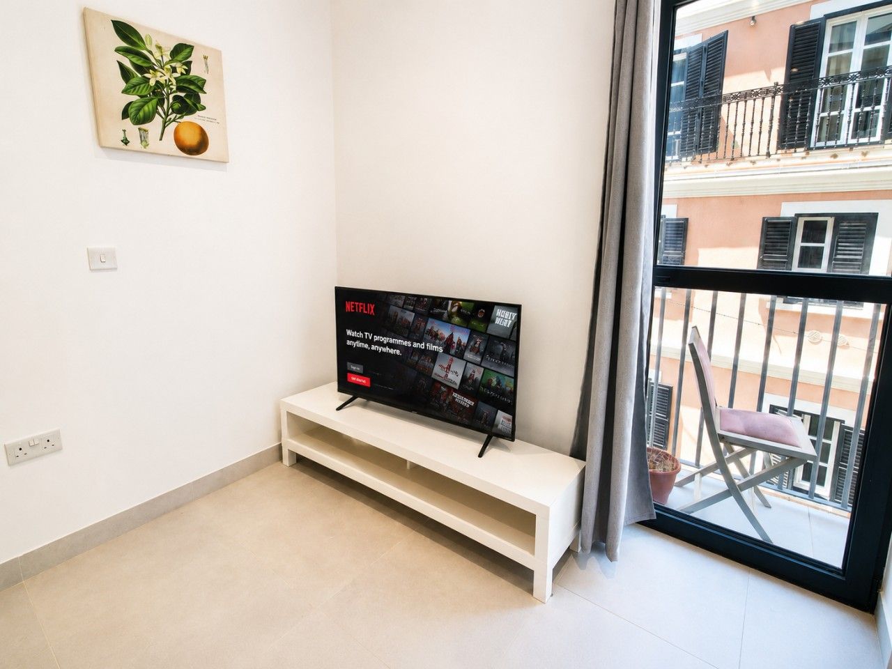A modern living room with a flat-screen TV on a white stand, a small white shelf, a potted plant, and a balcony view.