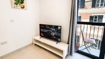 A modern living room with a flat-screen TV on a white stand, a small white shelf, a potted plant, and a balcony view.