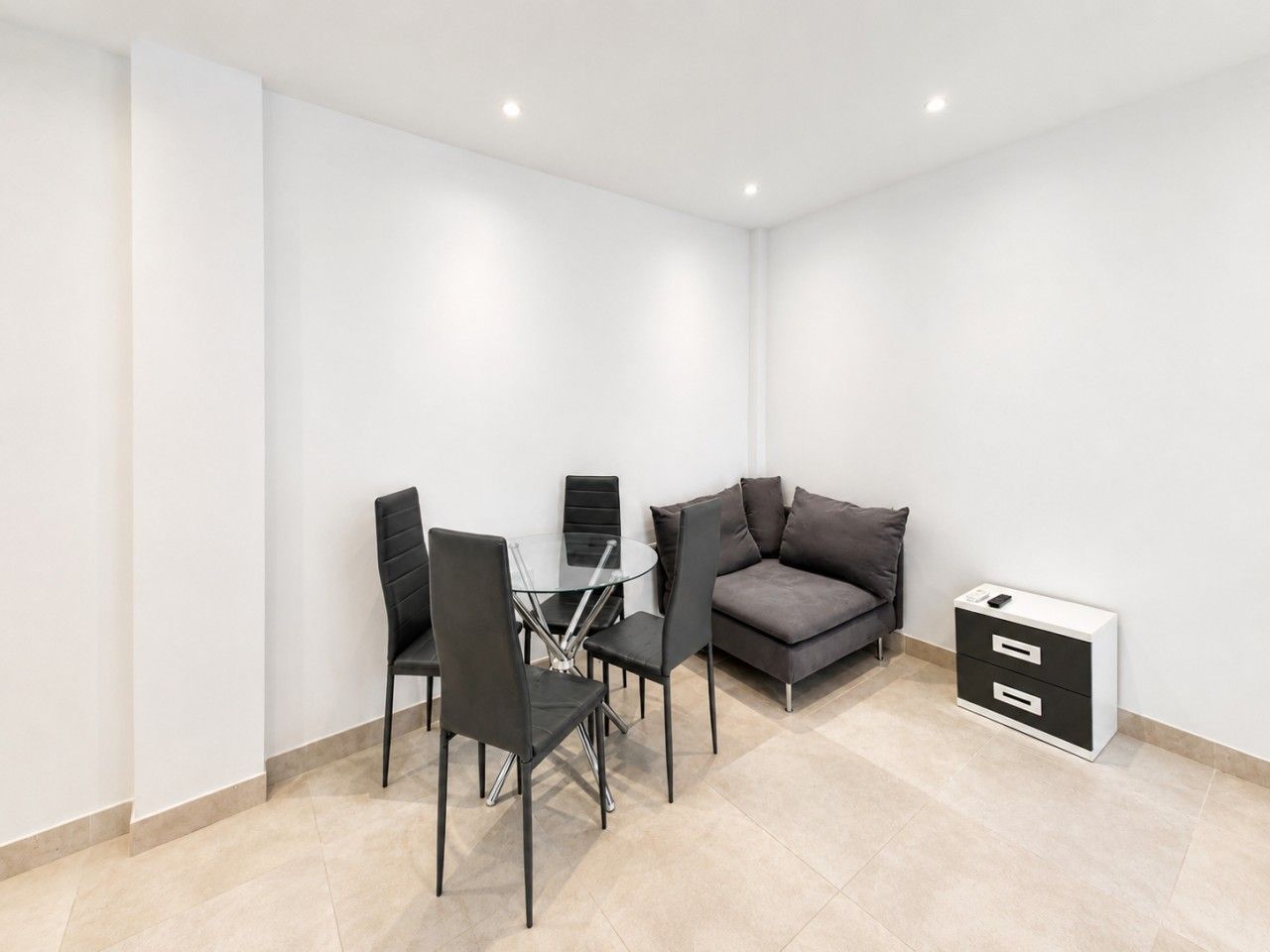 A modern room with a glass-top dining table, four chairs, a gray sofa, and a white storage unit.