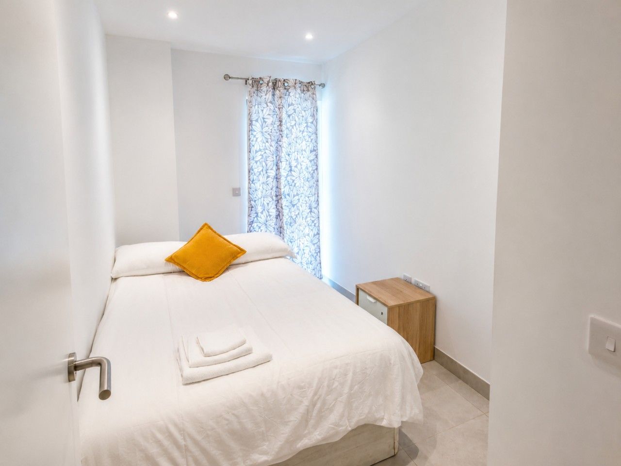 A hotel room with a bed, a nightstand, a window with curtains, and a door.