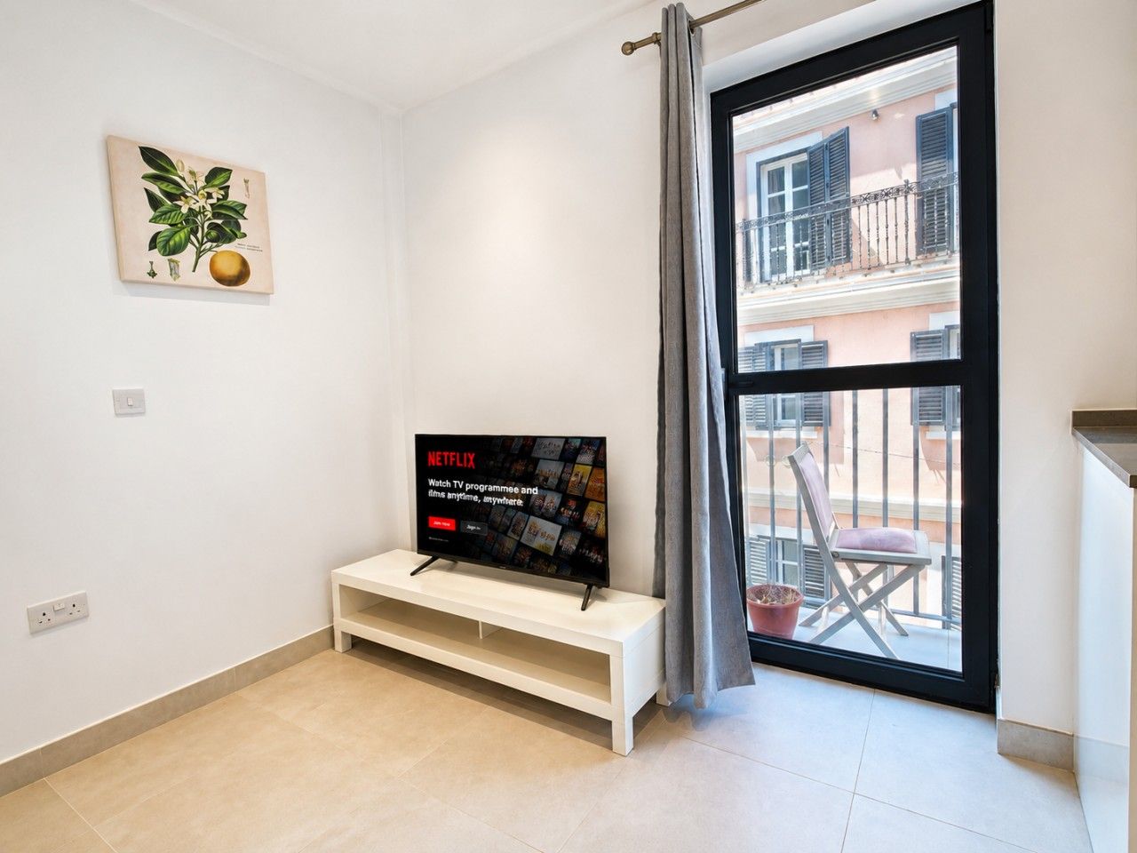 A modern living room with a flat-screen TV on a white stand, a potted plant on the wall, and a balcony with a chair and railing.