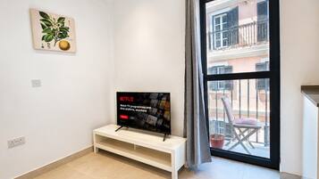 A modern living room with a flat-screen TV on a white stand, a potted plant on the wall, and a balcony with a chair and railing.