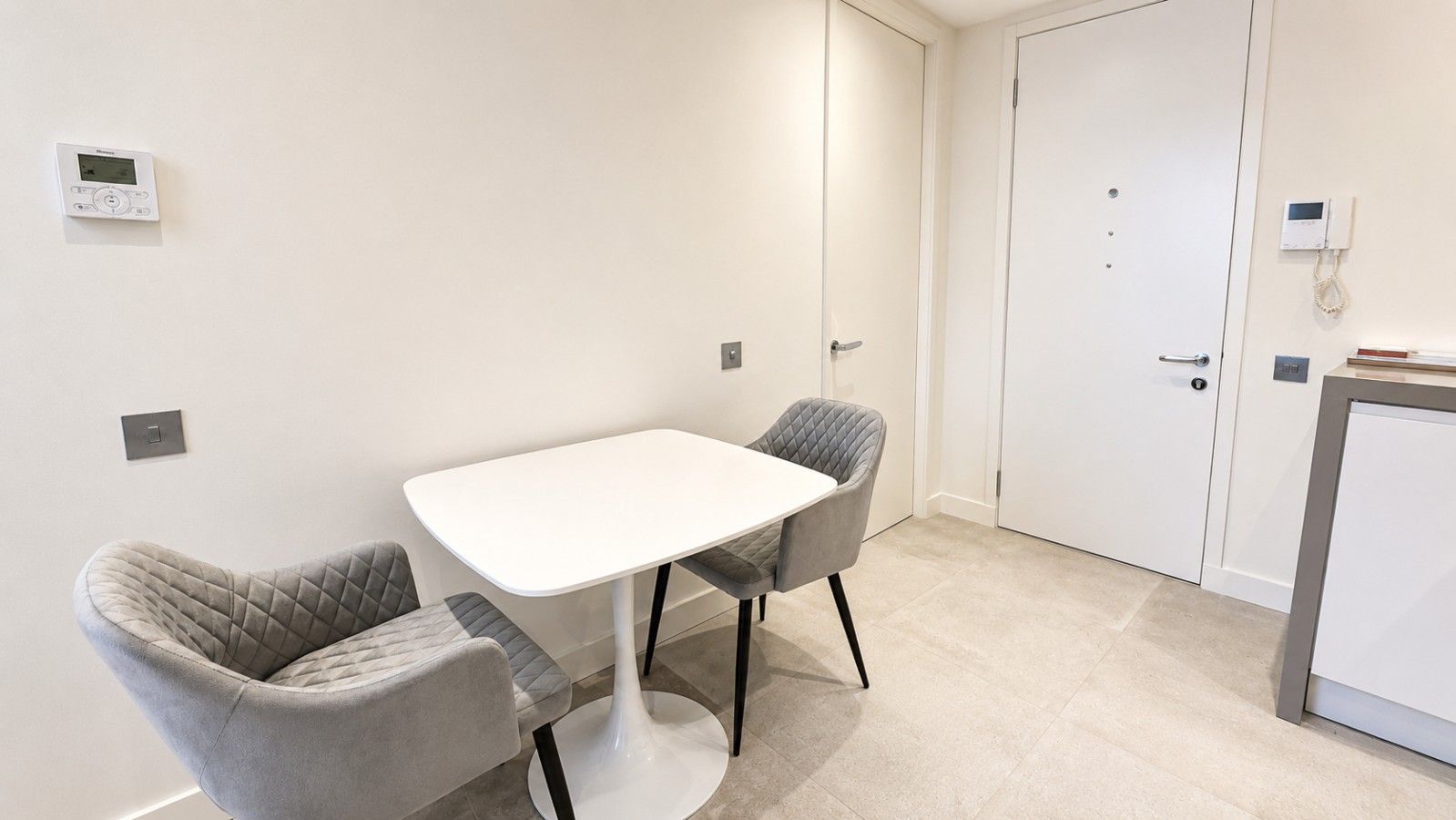 Een kleine, moderne kamer met een witte tafel en twee stoelen, een deur en een thermostaat aan de muur.