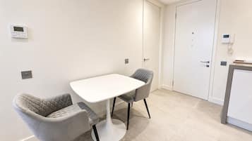 Een kleine, moderne kamer met een witte tafel en twee stoelen, een deur en een thermostaat aan de muur.