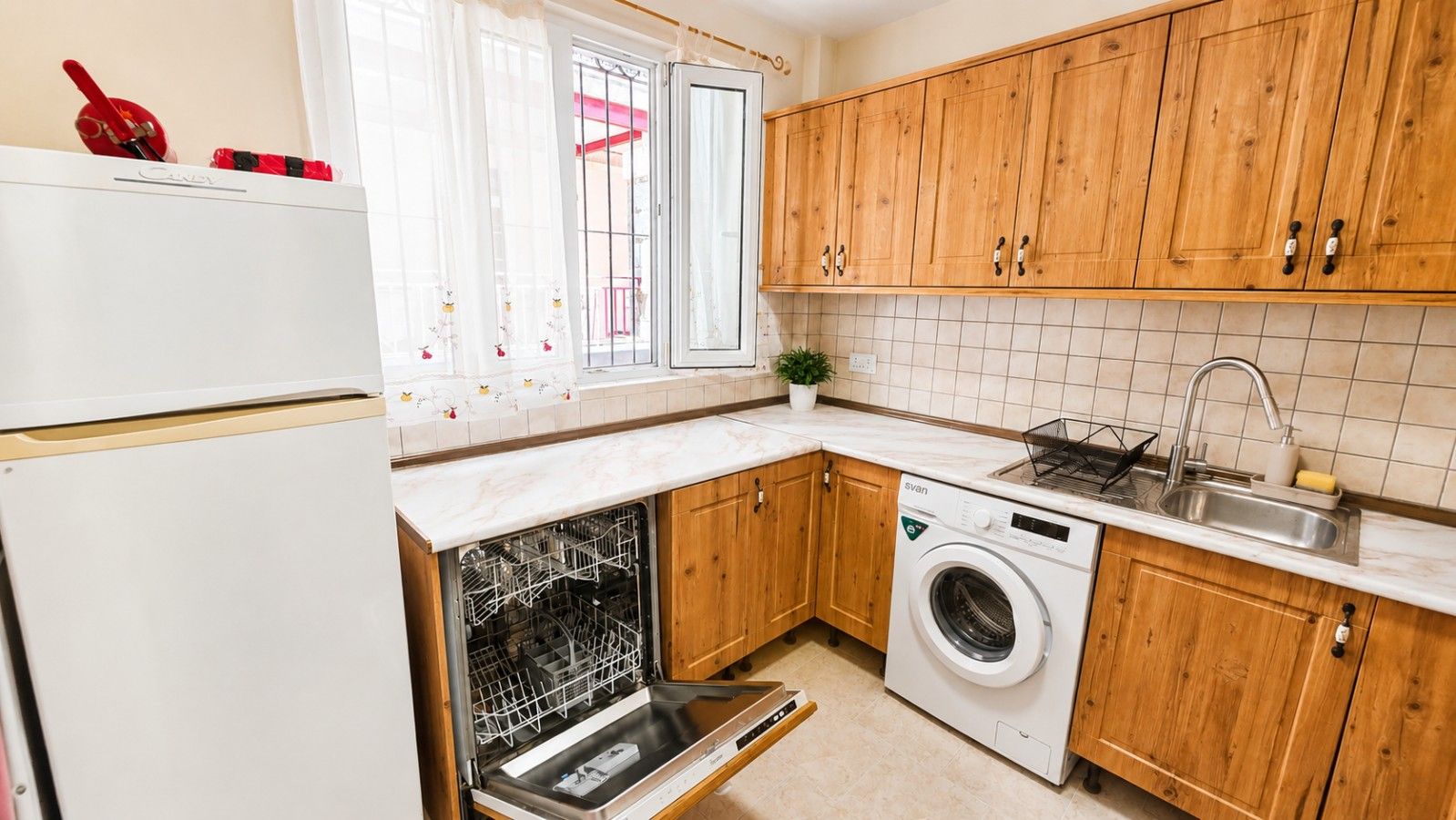 A compact kitchen with wooden cabinets, a built-in dishwasher, a washing machine, and a sink with a faucet.