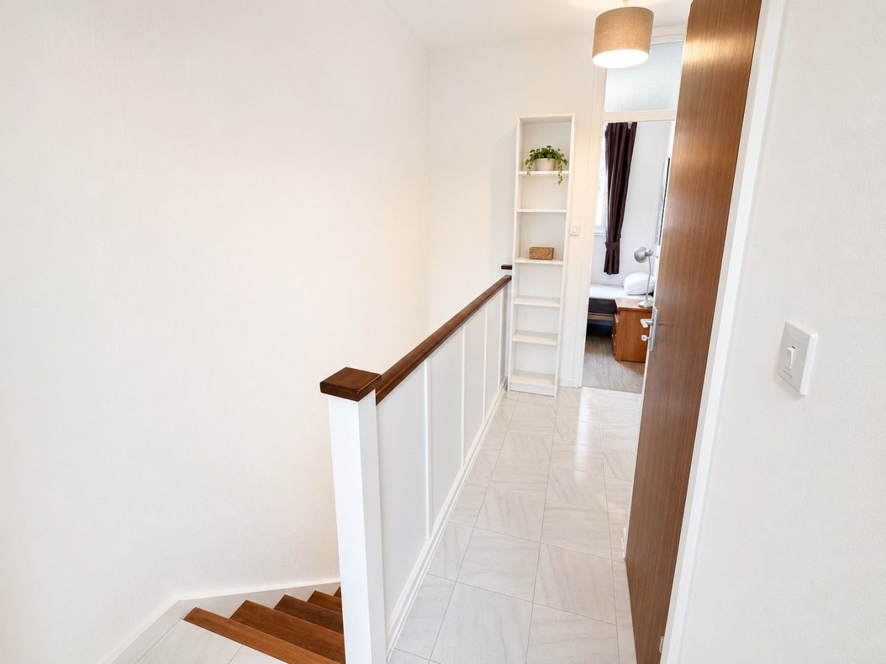A staircase with white walls and wooden handrails leading to a room with a bed, a nightstand, and a shelf with a plant.
