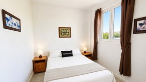 Quarto com cama, mesas de cabeceira, um criado-mudo, janela com cortinas e um quadro na parede.