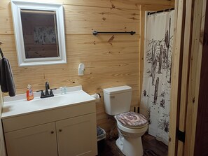A bathroom with a wooden wall, a white sink with soap and a toothbrush, a toilet with a decorative lid, and a shower curtain with a plant pattern. - The Yellow Rose- Lakehouse at Arc Ridge Ranch Resort (Ben Wheeler)