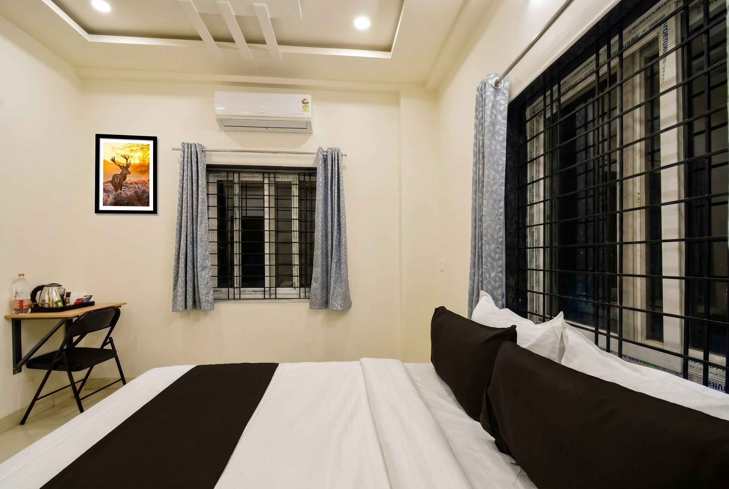 A hotel room with a bed, a desk, a chair, a window with metal bars, and an air conditioning unit.