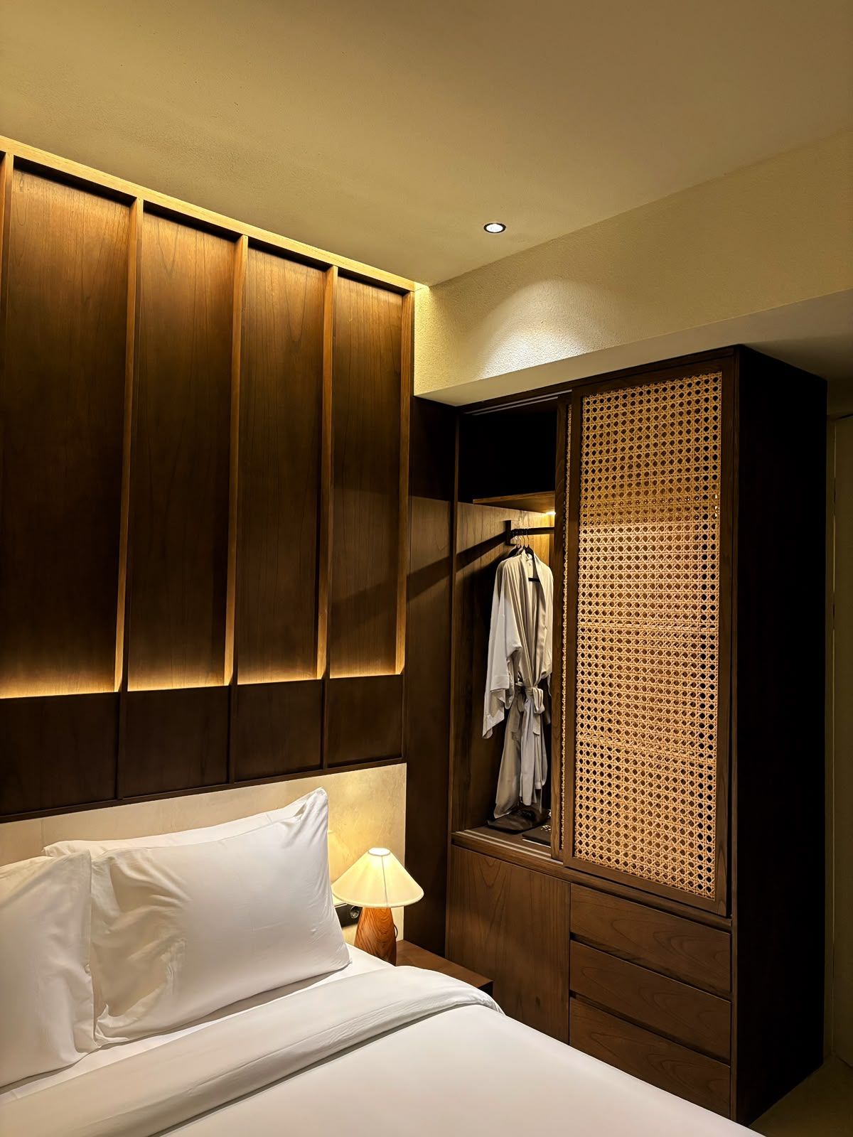 Habitación de hotel con cama, mesita de noche, lámpara, armario y bata colgada.