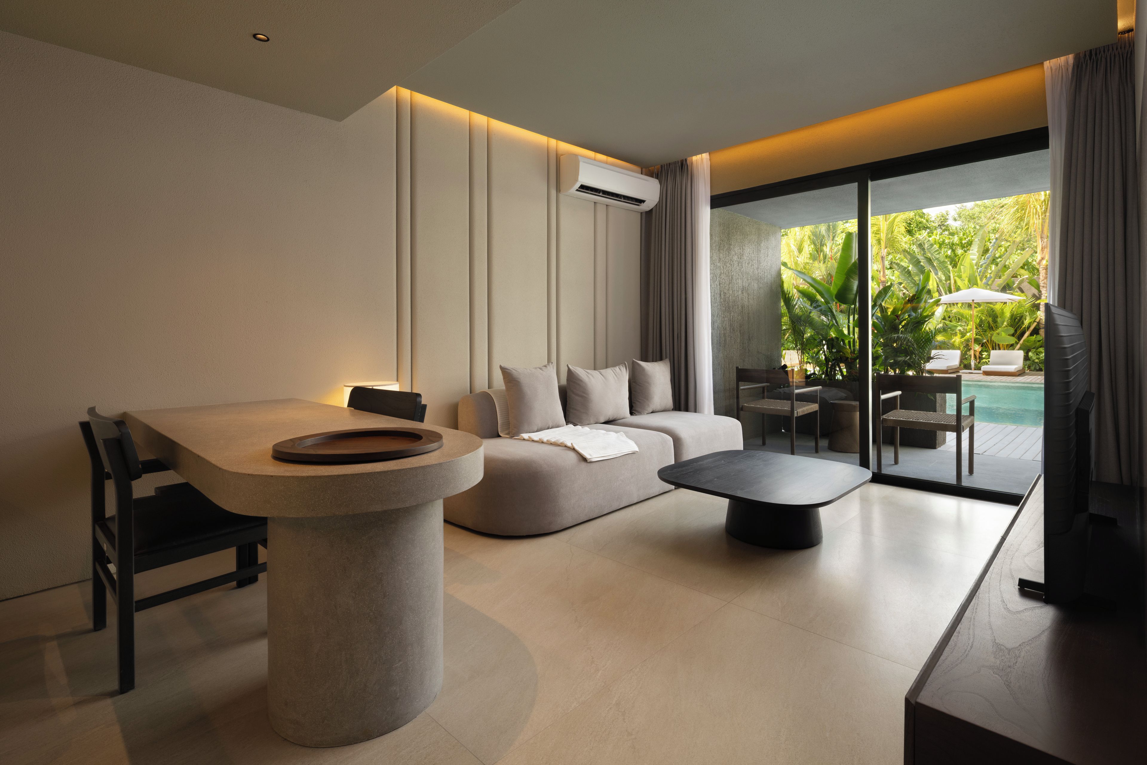 A modern living room with a dining area, a sofa, a coffee table, and a view of a pool and greenery.