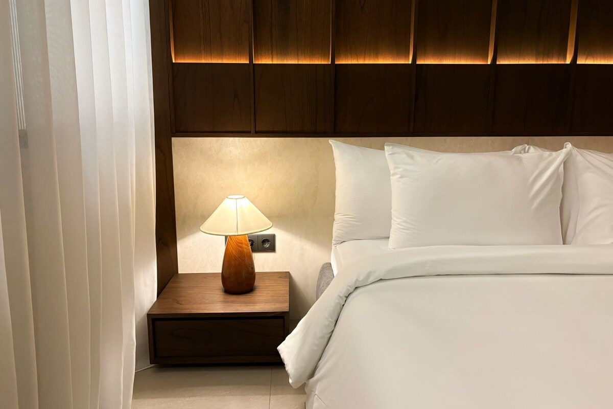 A hotel room with a wooden headboard, a bedside table with a lamp, and a bed with white bedding.