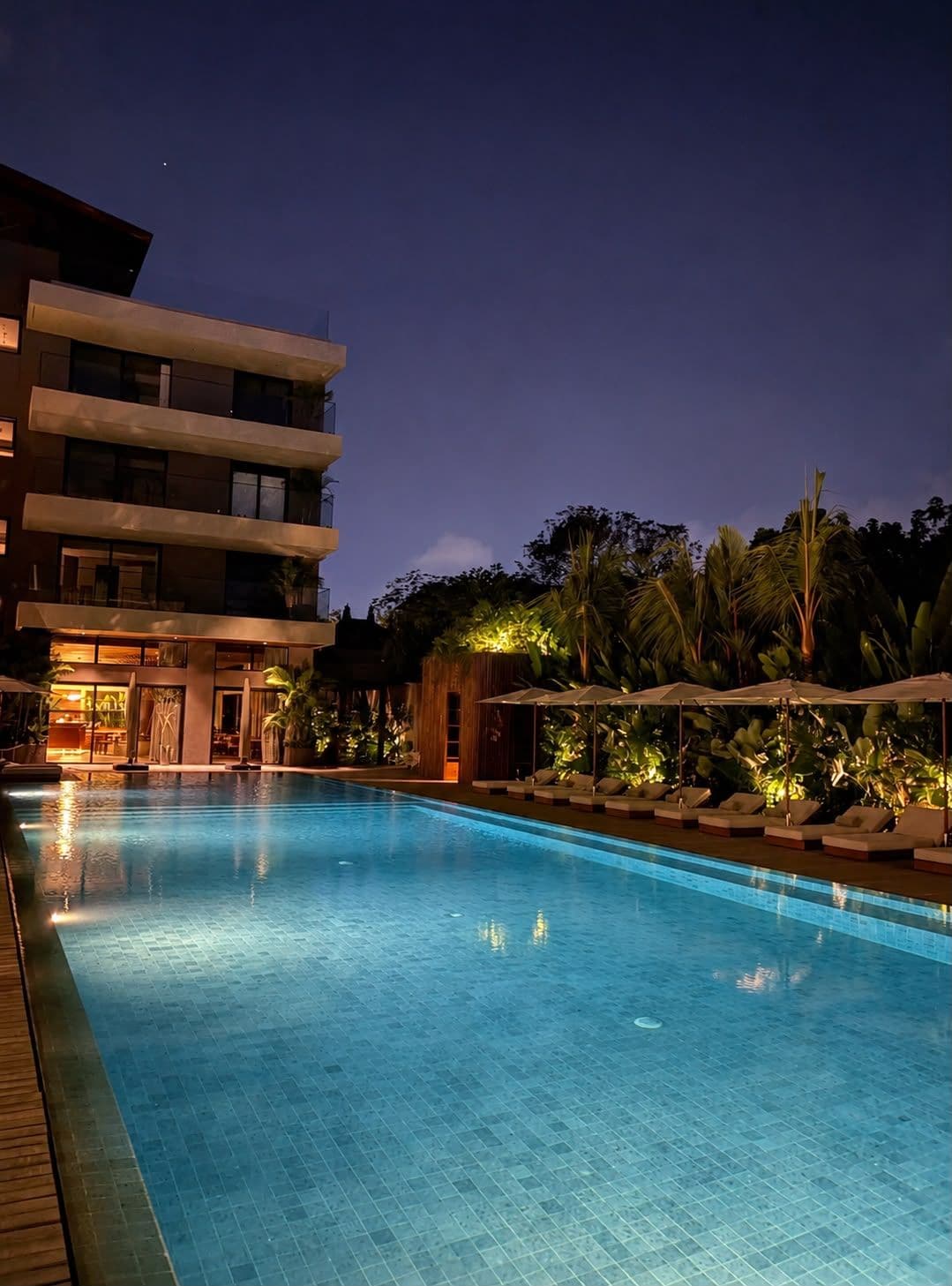Piscina con sillones de descanso y sombrillas, rodeada por un edificio de varios pisos y vegetación tropical.