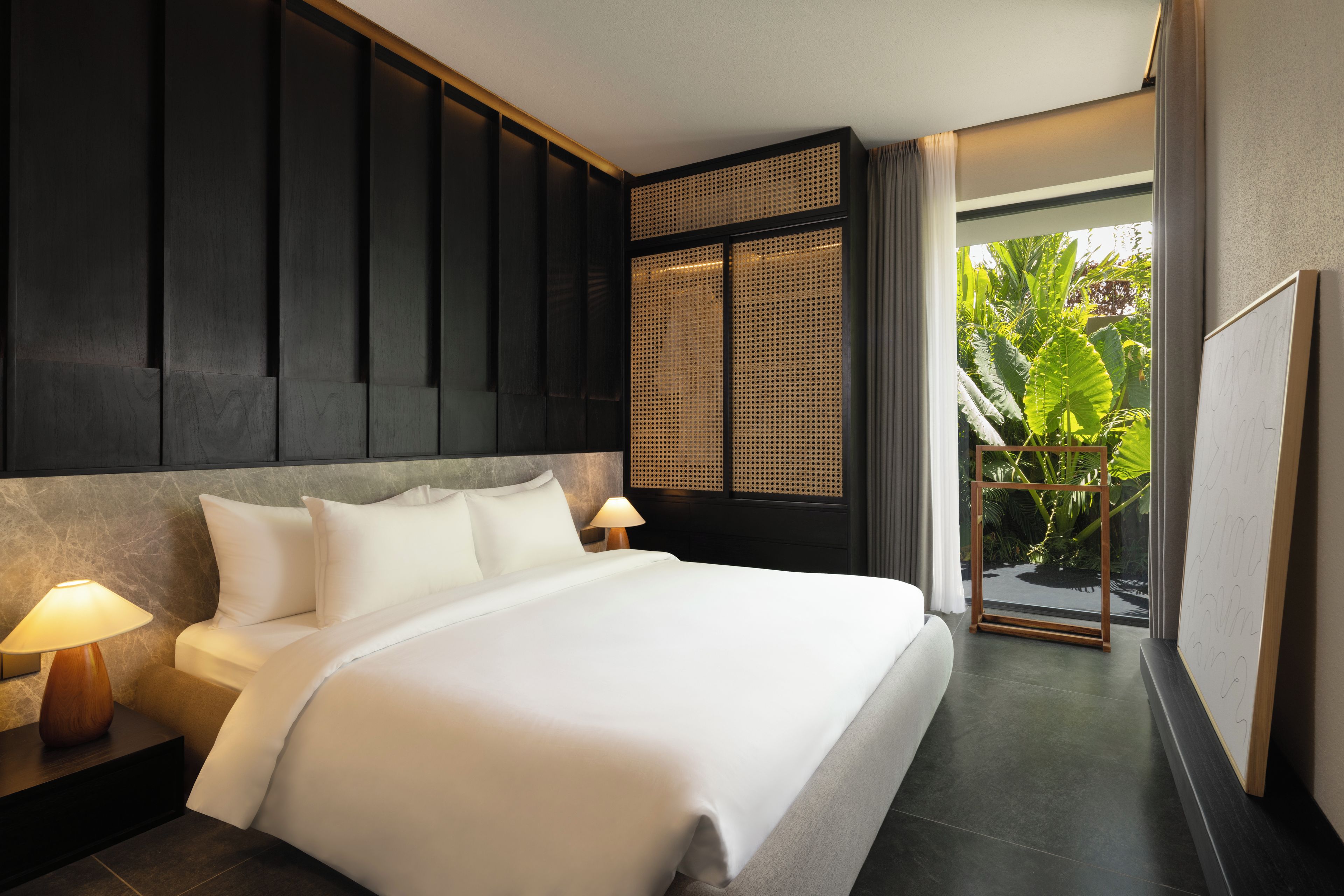 Habitación de hotel moderna con una cama grande, lámparas de noche, vistas a la vegetación por una ventana y un panel tejido tradicional.