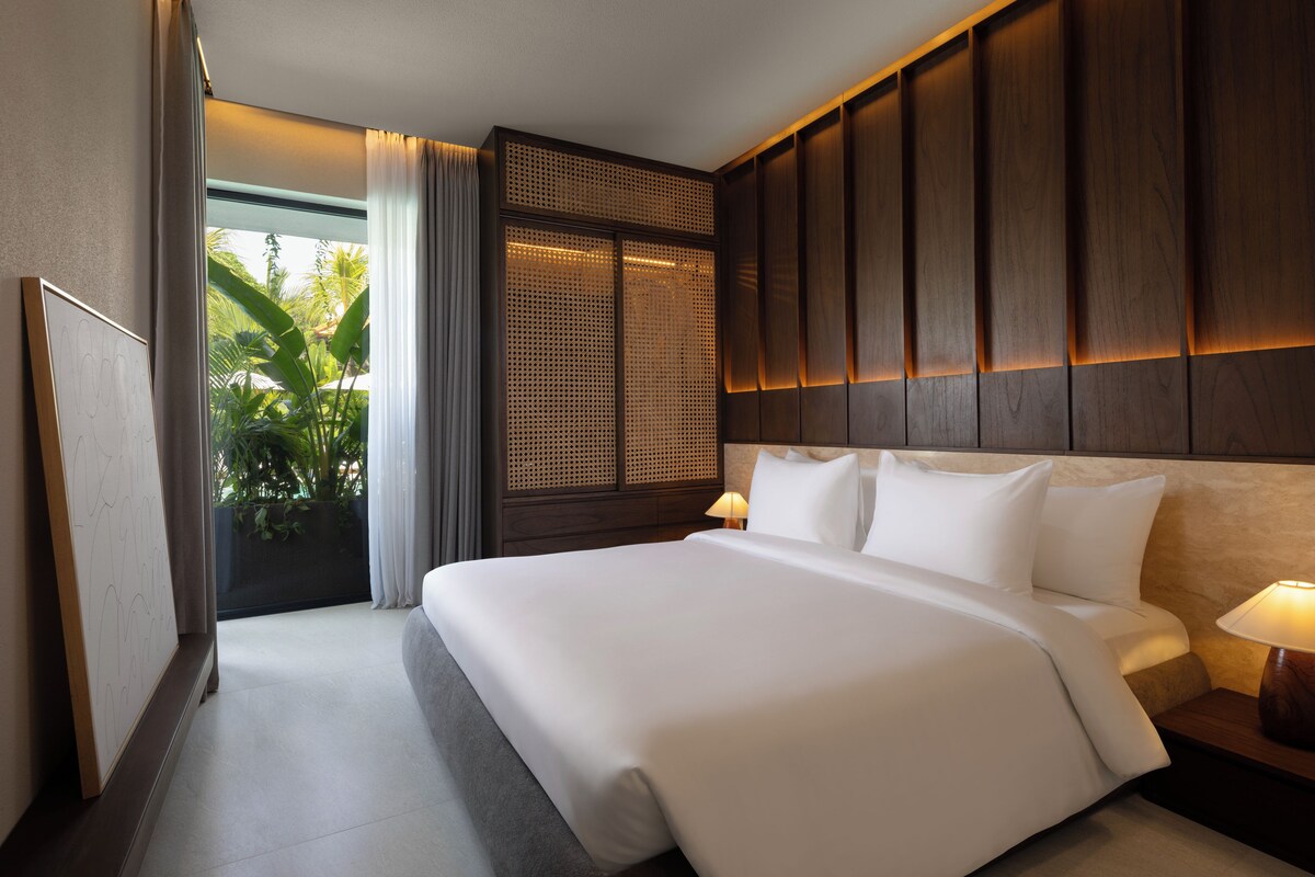 Habitación de hotel con una cama grande, una mesita de noche, una lámpara, una ventana con cortinas y vistas al exterior.