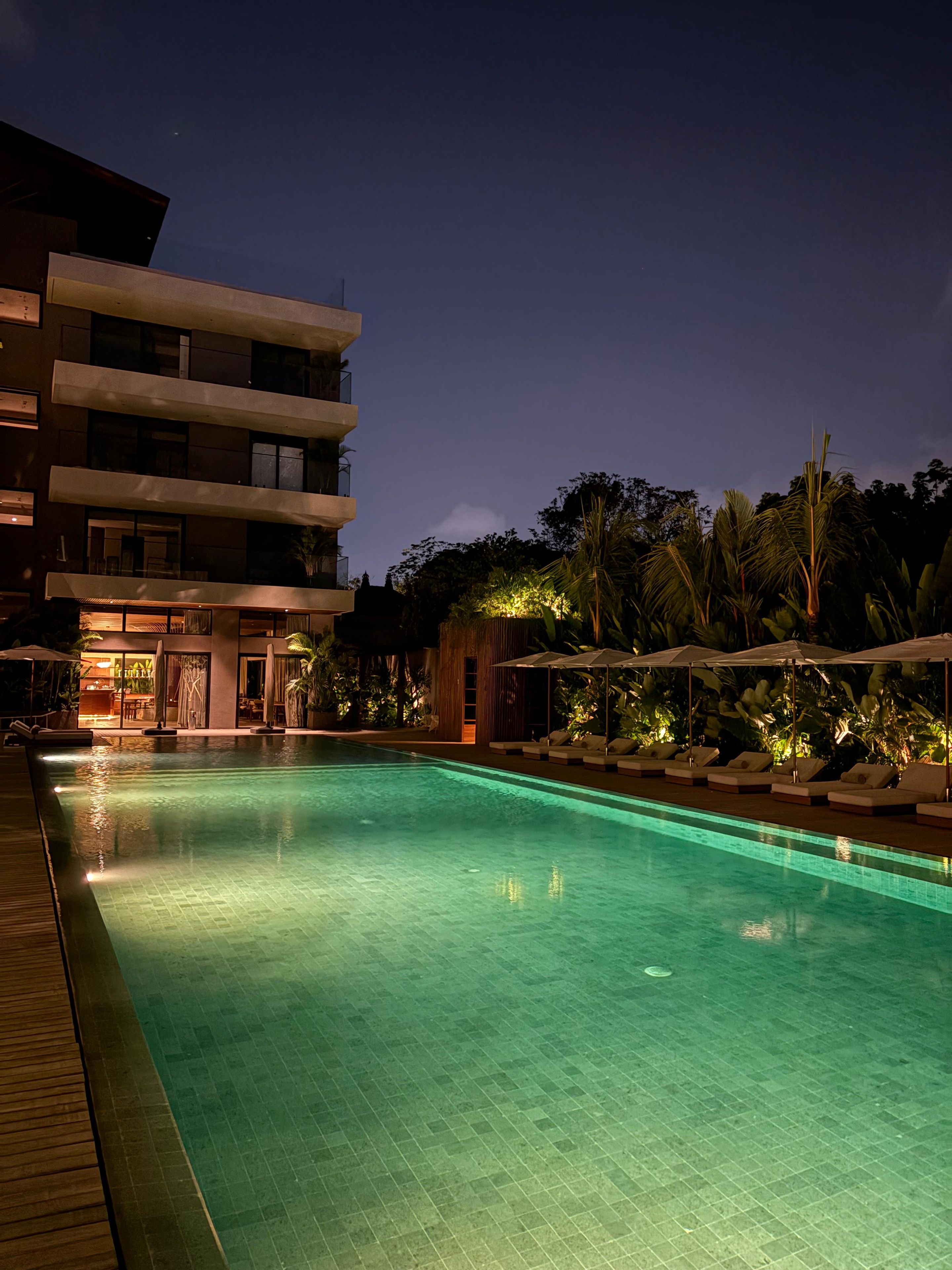 A nighttime swimming pool with a wooden deck, lounge chairs, and a building with multiple floors and balconies.