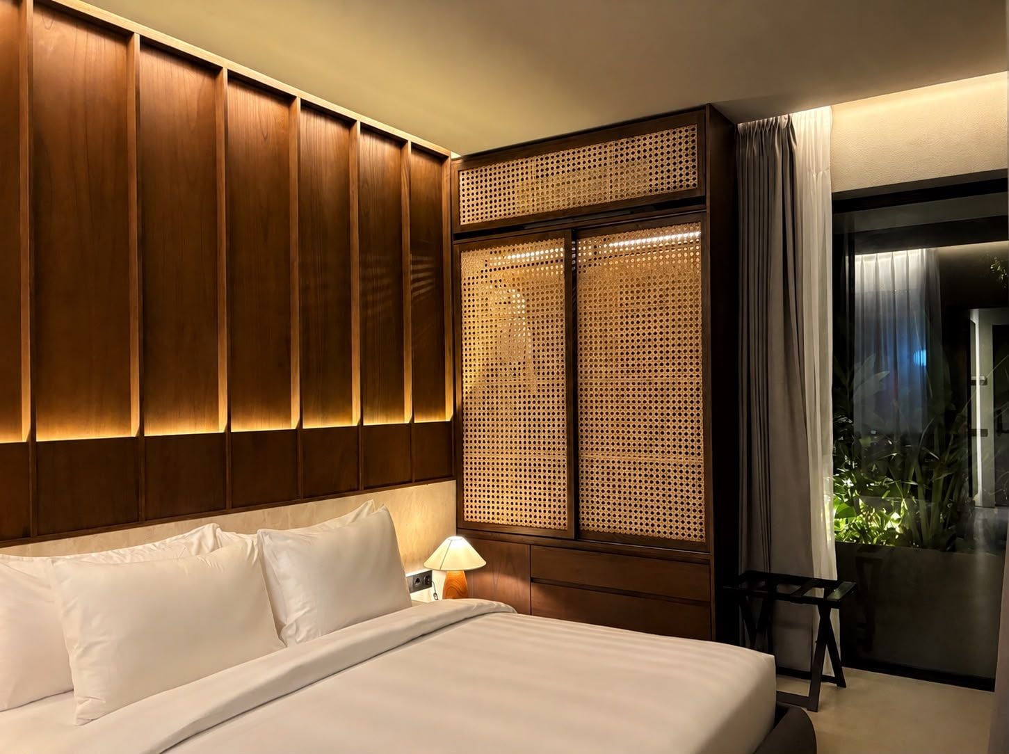 A hotel room with a large bed, wooden paneling, a woven partition, a nightstand with a lamp, and a view of greenery through a window.