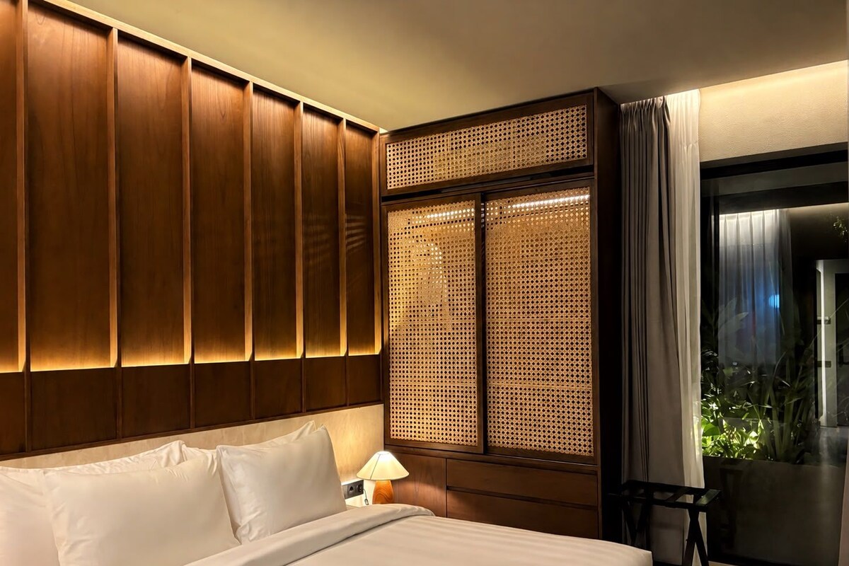 A hotel room with a large bed, wooden paneling, a woven partition, a nightstand with a lamp, and a view of greenery through a window.