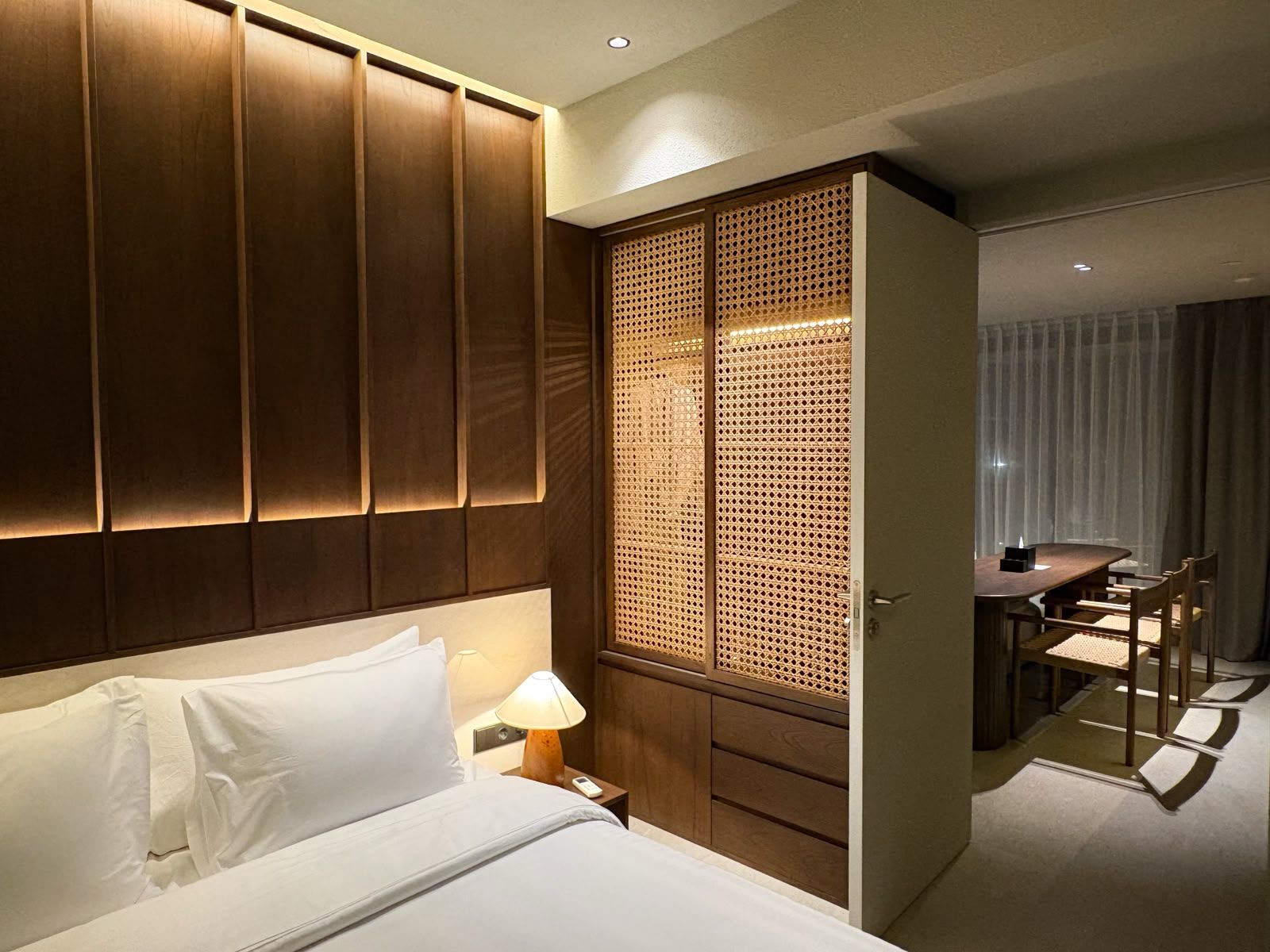 A hotel room with a bed, bedside table, lamp, wooden wardrobe, and a dining area with a table and chairs.