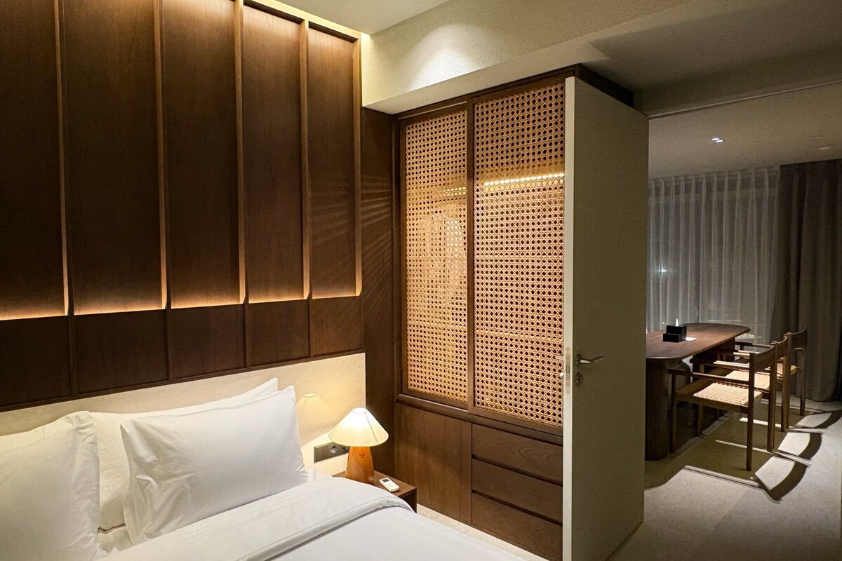 A hotel room with a bed, bedside table, lamp, wooden wardrobe, and a dining area with a table and chairs.