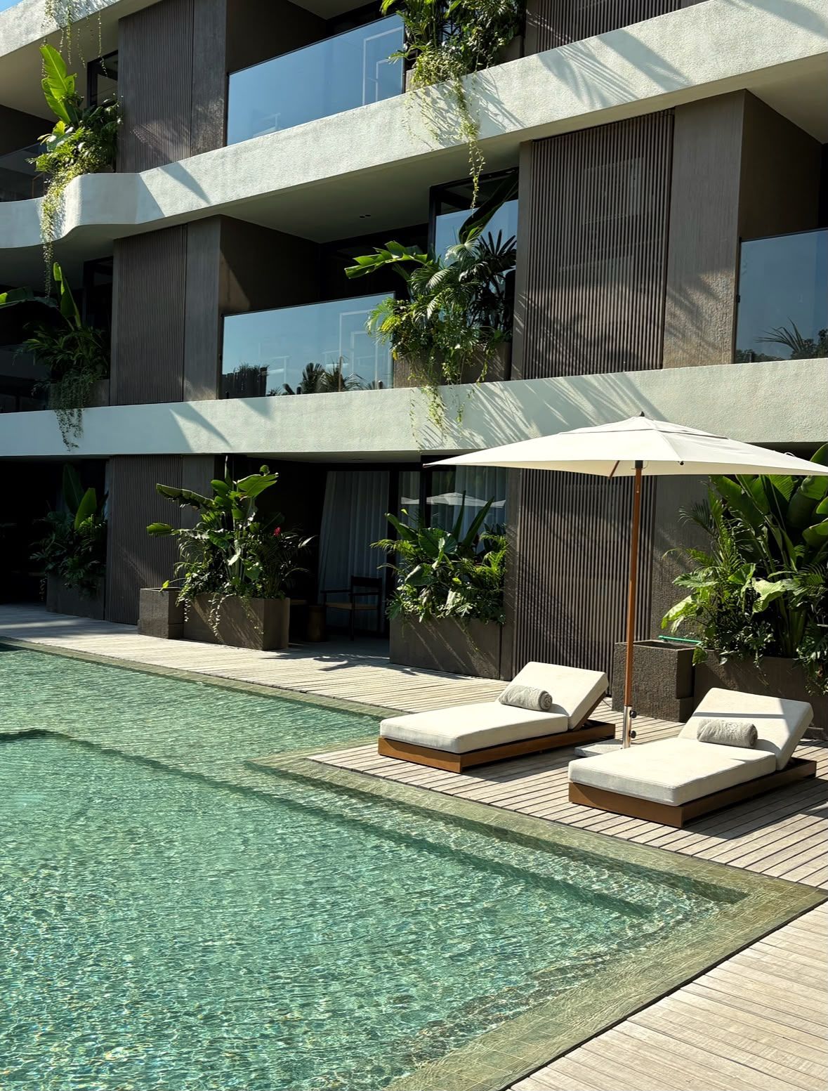 A modern hotel pool area with lounge chairs and an umbrella, surrounded by greenery and a multi-story building.
