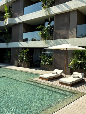 A modern hotel pool area with lounge chairs and an umbrella, surrounded by greenery and a multi-story building. - Elison Hotel Bali (Tibubeneng)