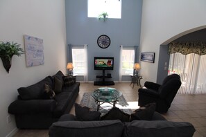 Ein Wohnzimmer mit einer Couch, einem Sessel, einem Glastisch und einem Fernseher. An der Wand hängt eine Uhr und ein Fenster mit Jalousien.