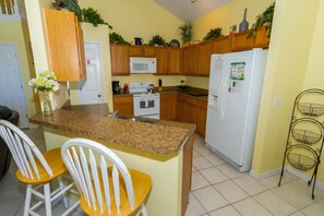 A kitchen with wooden cabinets, a white refrigerator, a microwave, and a stove. There are two white chairs and a small table. - Home With a View! (378106) (Kissimmee)