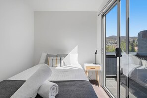 A modern bedroom with a large bed, a bedside table, a lamp, and a view of a cityscape through a glass door. - Warwick 1 – Stylish Hobart Retreat (Hobart)