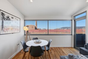 A modern room with a dining table, chairs, a floor lamp, and large windows offering a view of buildings and a red-roofed structure. - Warwick 1 – Stylish Hobart Retreat (Hobart)