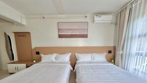 Hotellihuone, jossa on kaksi sänkyä, puinen pääty, pieni yöpöytä, jossa on kasvi, seinään asennettu ilmastointilaite ja ikkuna, jossa on verhot.