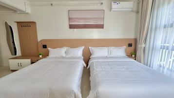 A hotel room with two beds, a wooden headboard, a small bedside table with a plant, a wall-mounted air conditioner, and a window with curtains.