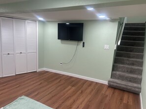 Una habitación con piso de madera, un televisor montado en la pared, una escalera y un armario blanco.