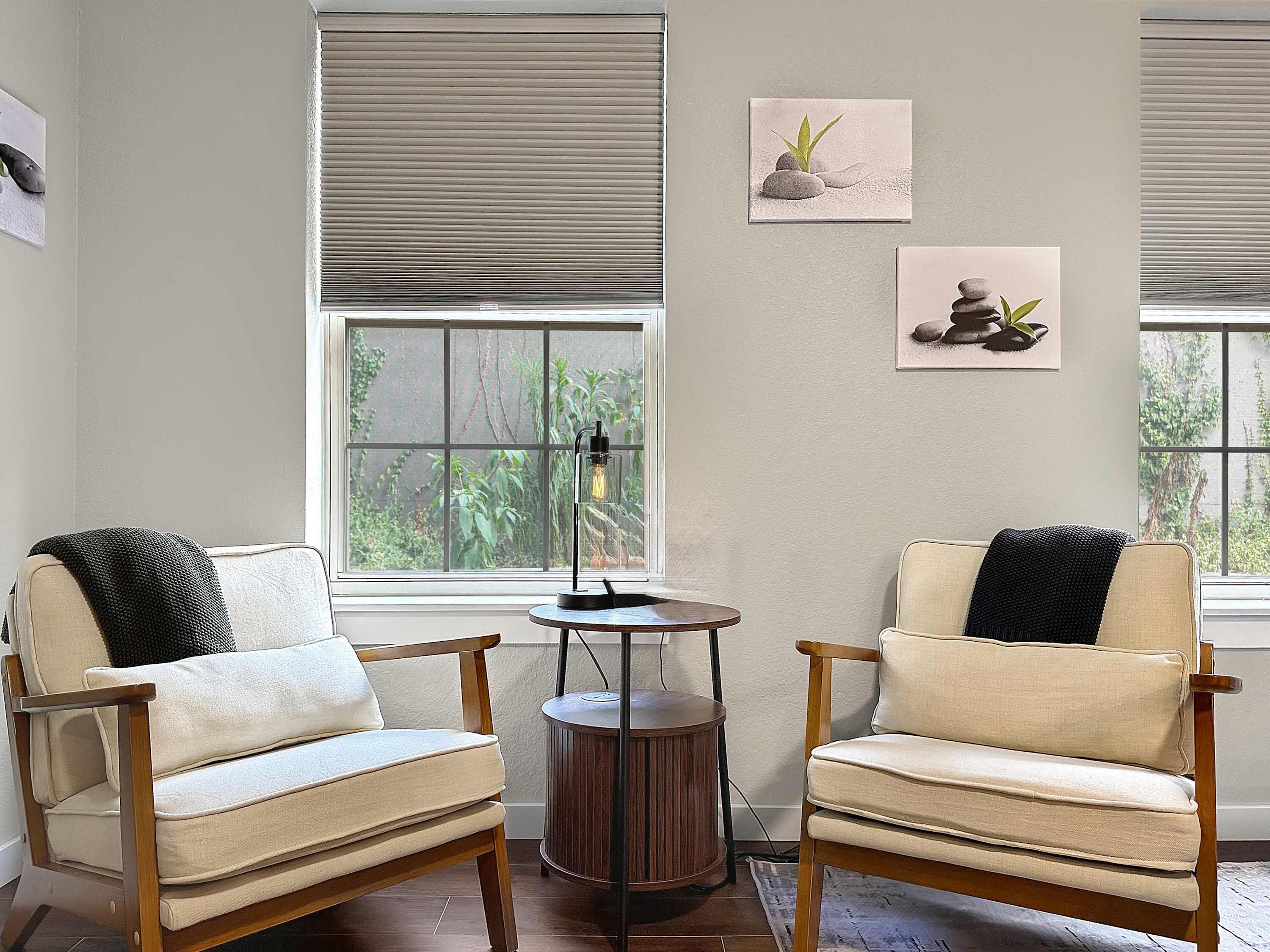 A room with two beige armchairs, a small wooden table, and a floor lamp. There are two windows with blinds, and artwork on the walls.