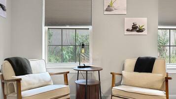 A room with two beige armchairs, a small wooden table, and a floor lamp. There are two windows with blinds, and artwork on the walls.