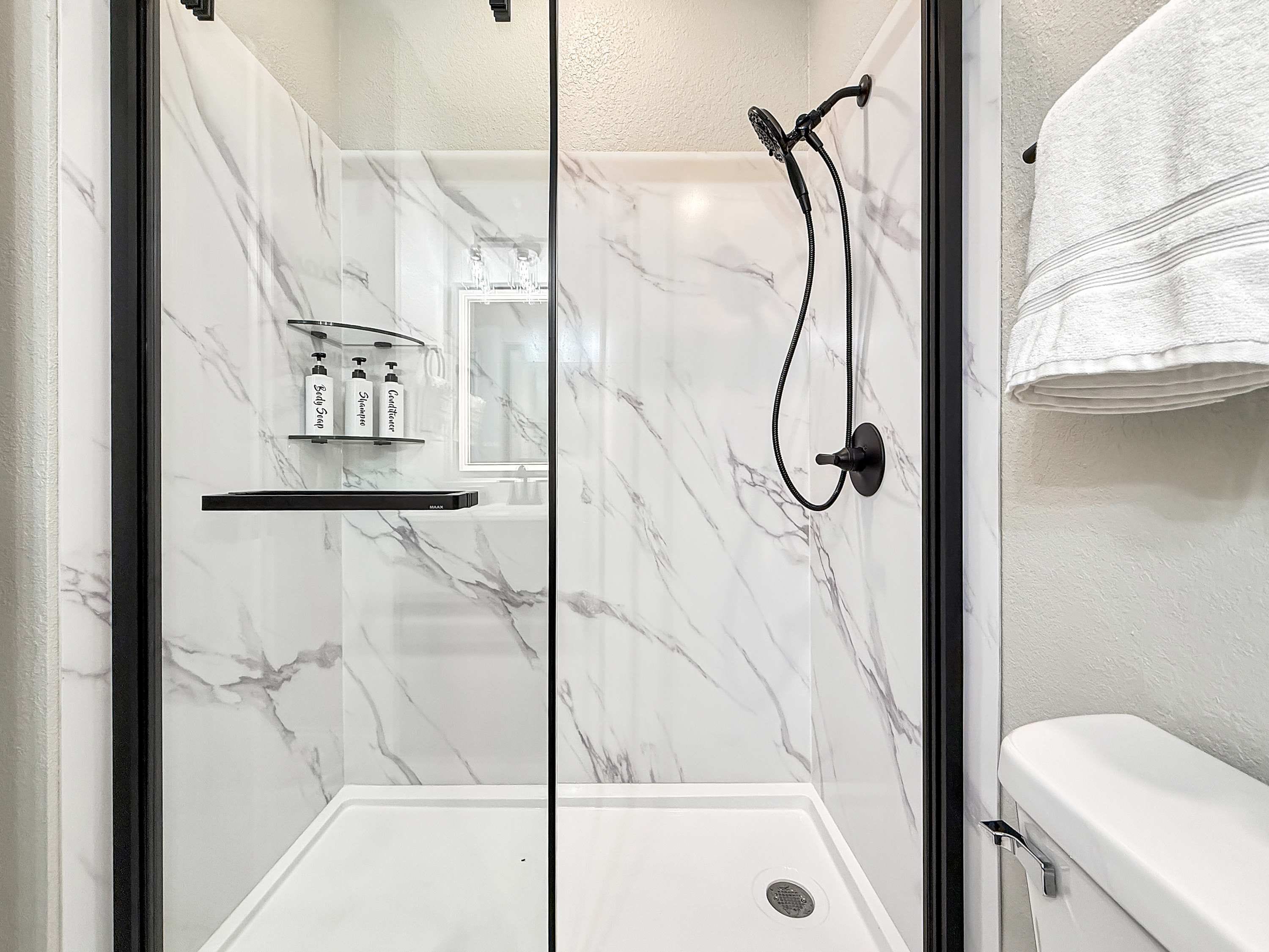 A glass-enclosed shower with a handheld showerhead, a shelf with toiletries, and a white towel hanging on the wall.