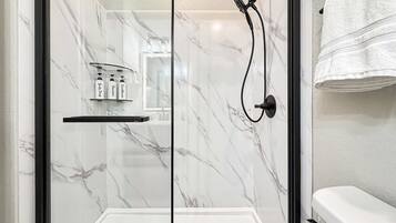 A glass-enclosed shower with a handheld showerhead, a shelf with toiletries, and a white towel hanging on the wall.