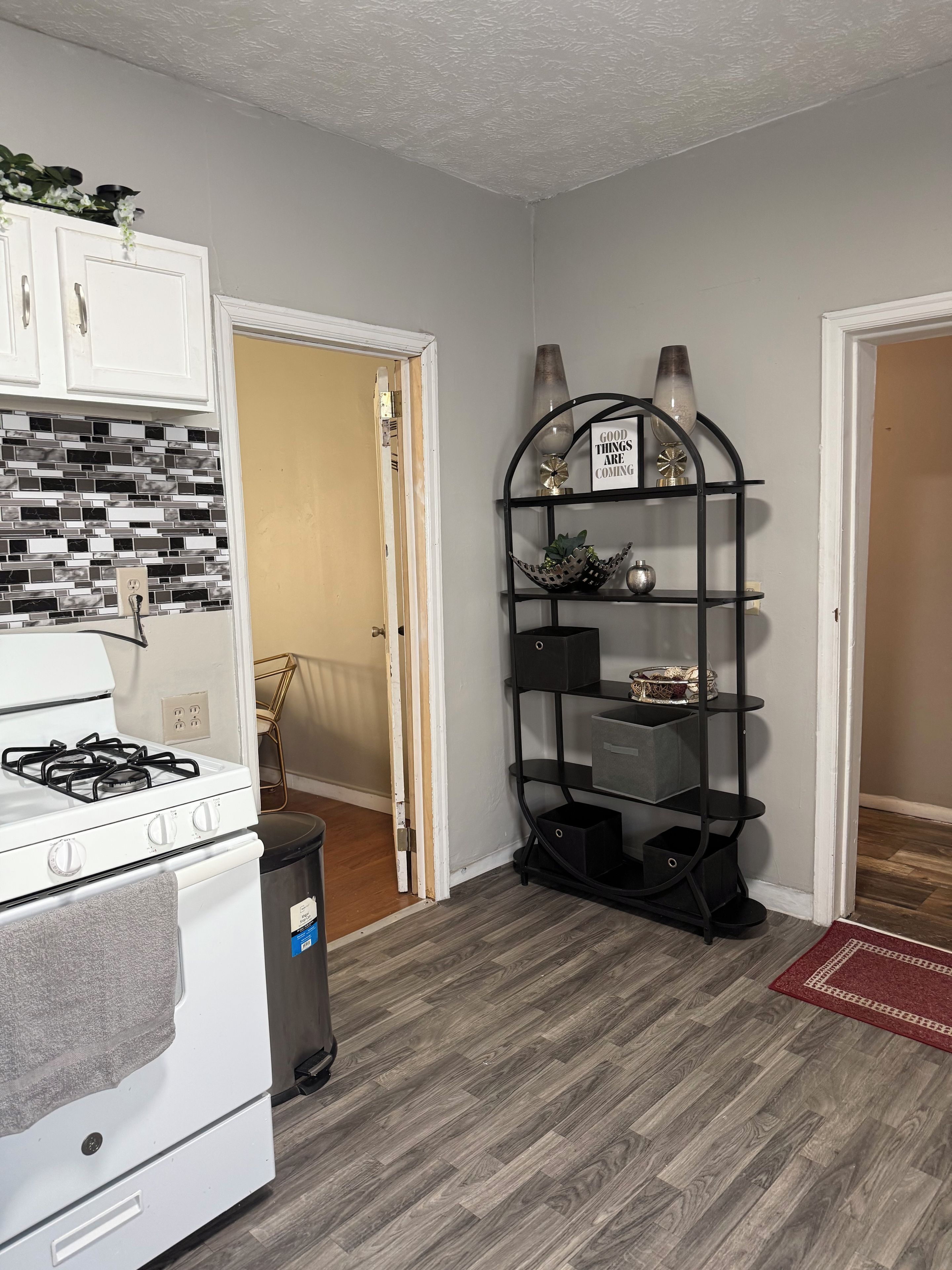 A kitchen with a white stove, a trash bin, and a black metal shelf unit with decorative items.