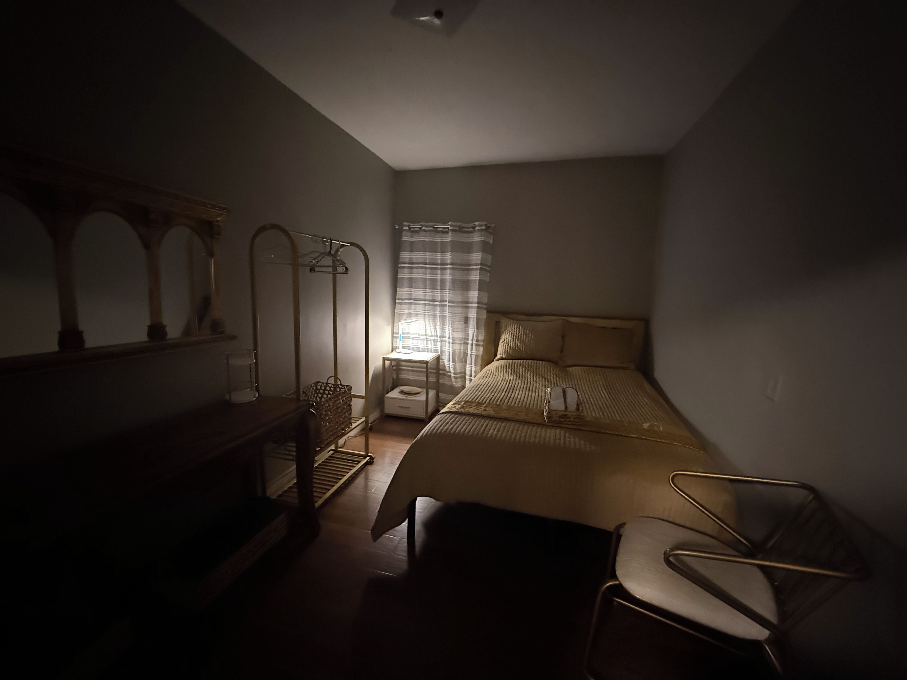 A dimly lit bedroom with a bed, a chair, a mirror, and a nightstand.