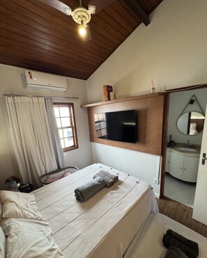 Quarto com cama, TV fixada na parede, ventilador de teto e banheiro visível por uma porta aberta.