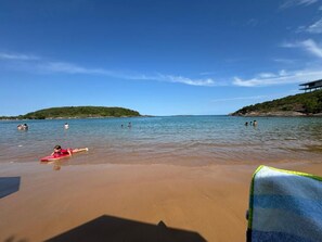 Uma praia com águas azuis cristalinas, uma pessoa de fato de banho vermelho e uma cadeira de praia verde e azul.