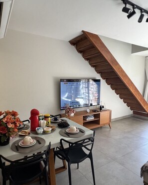 Uma área de refeições com uma mesa posta para dois, uma televisão fixada na parede e uma escada de madeira.